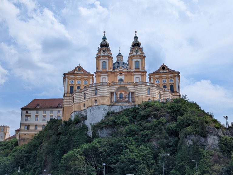 Melk abbey from a street far below. Green trees in front of the abbey set on a hill. Blue sky with many big clouds.