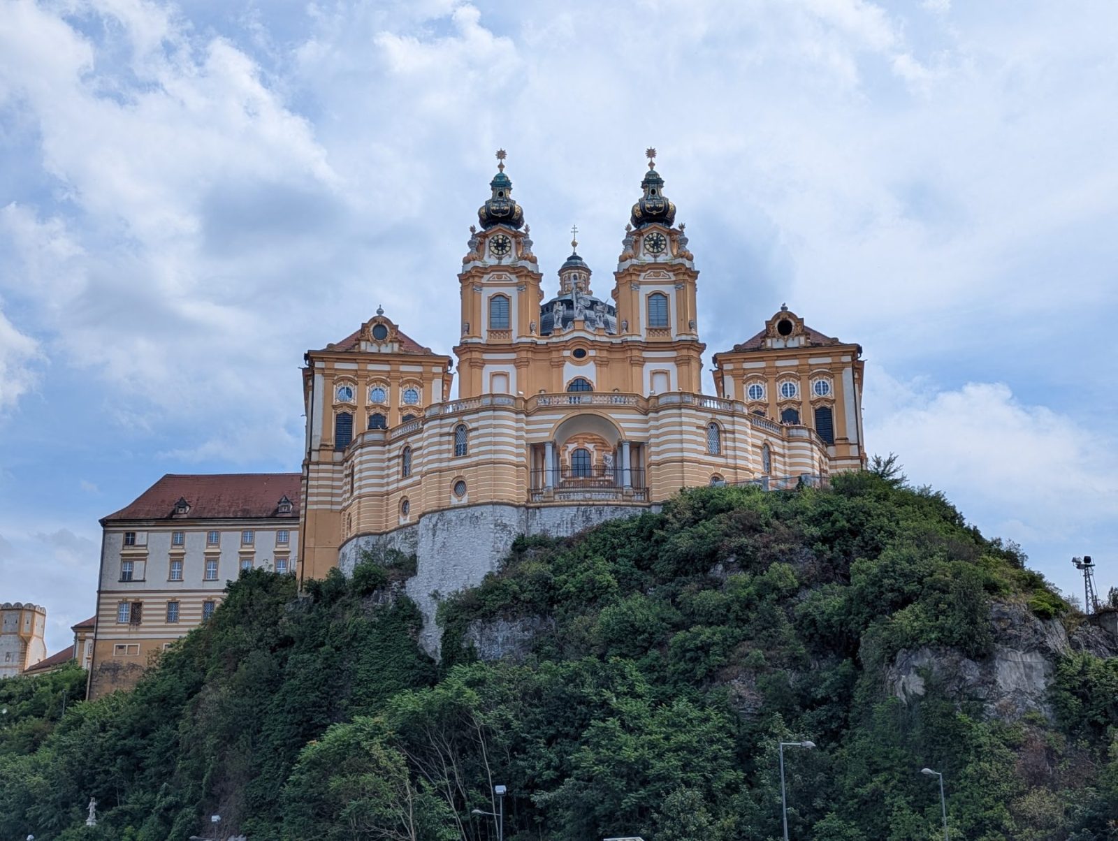 Melk abbey from a street far below. Green trees in front of the abbey set on a hill. Blue sky with many big clouds.