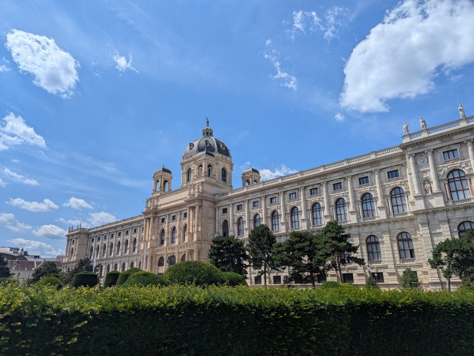 Historic museum building in Vienna, Austria, seen from a garden with green hedges under a bright blue sky with clouds.