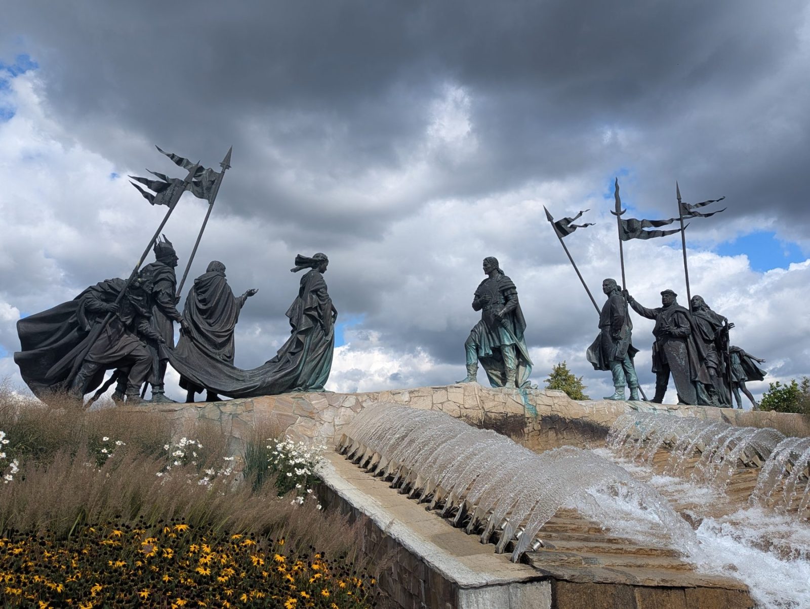he Nibelungen statue in Tulln, Austria, showing bronze warrior figures with flags standing above a cascading fountain and wildflowers on a cloudy day.