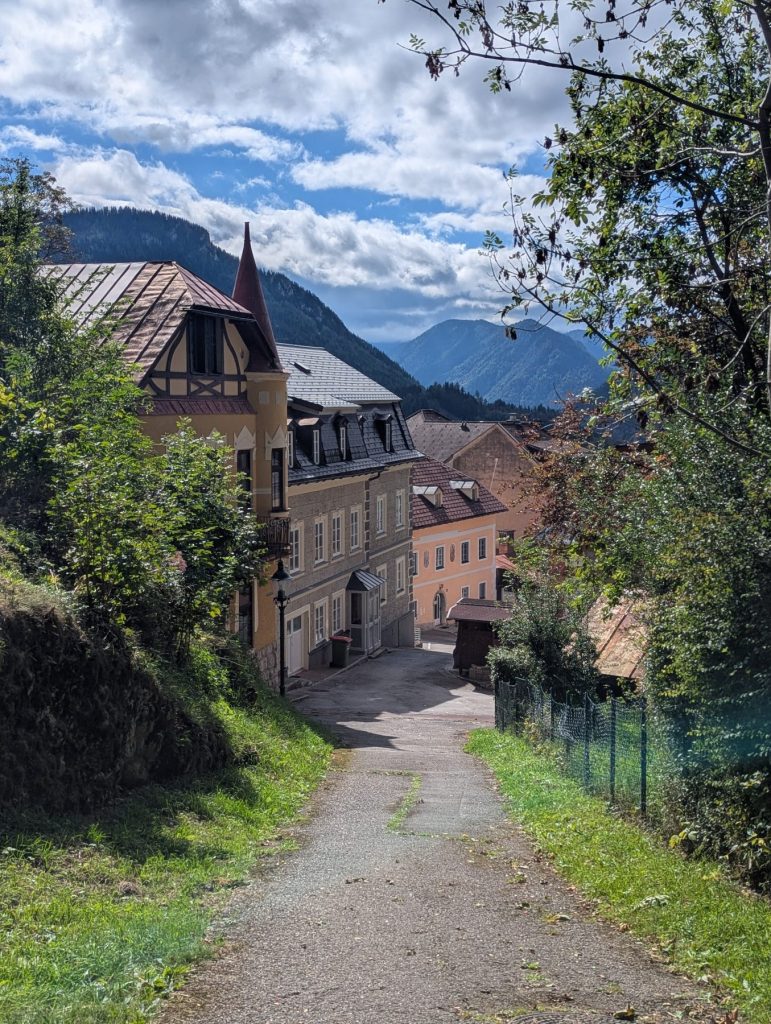 The TIST Assistant said: Sunlit path leading downhill to colourful historic houses in Mariazell, framed by green trees with misty Austrian mountains and a partly cloudy sky in the background.