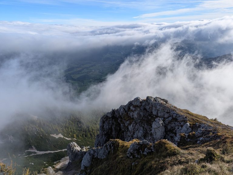 View from the Schneeberg summit in Lower Austria, showing a rocky outcrop above misty clouds and green valleys hidden below.