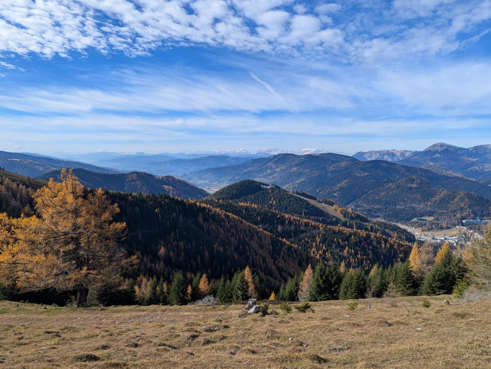 View from a mountain onto mountain range coloured in autumn colours. Green and orange trees scattered before blue sky.