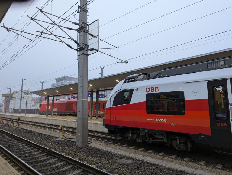 Two OEBB trains waiting at a train station. An empty train track in the foreground. Overcast sky.