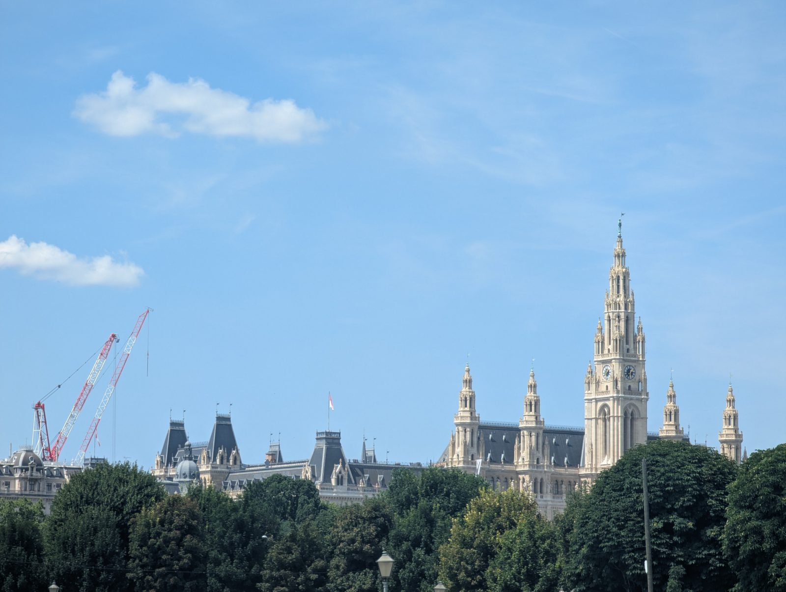 Vienna City Hall (Rathaus) rising above green trees, with its tall neo-Gothic clock tower and ornate spires set against a bright blue sky.