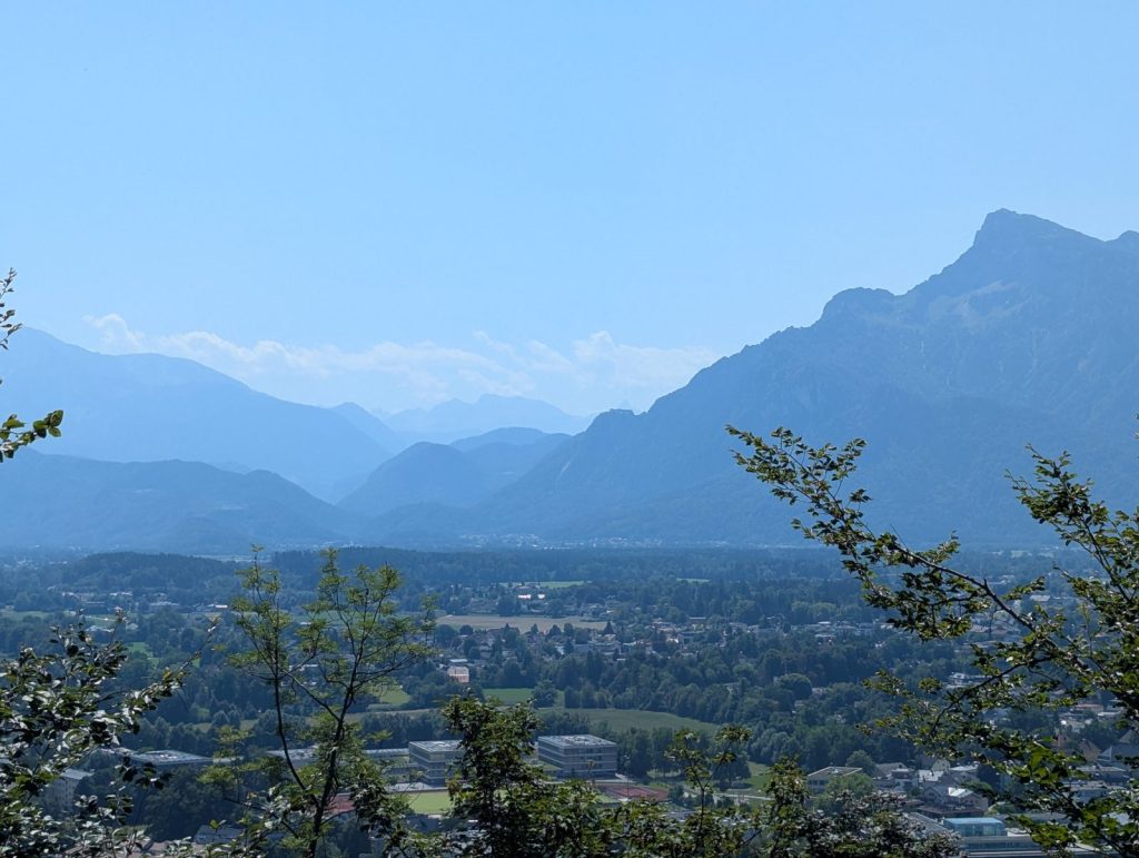 Panoramic view from Kapuzinerberg in Salzburg, looking out over the city and surrounding valley towards layered Alpine mountains under a clear blue sky.