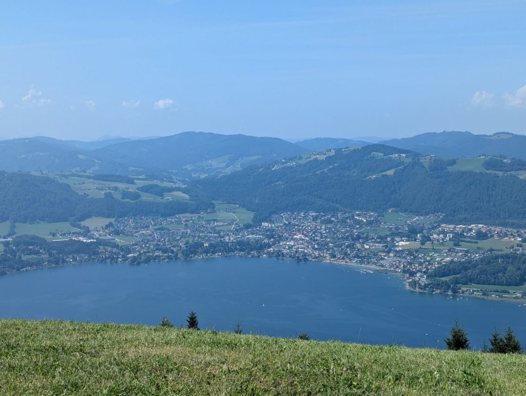 Panoramic view over Lake Traunsee in Gmunden, with the town along the shoreline and forested hills rising into the distance under a clear blue sky.