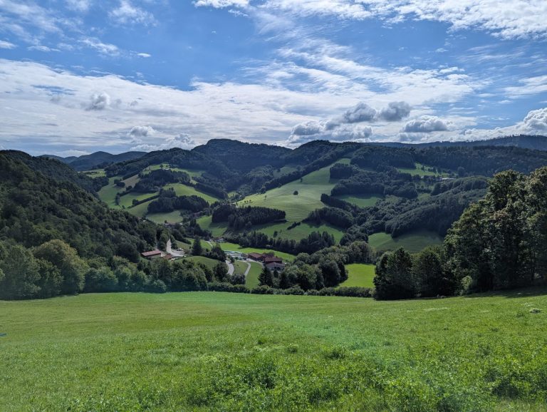 Wide view over the rolling green hills of the Pielachtal, with scattered farmhouses, winding roads, and forested slopes under a dramatic blue sky with layered clouds.