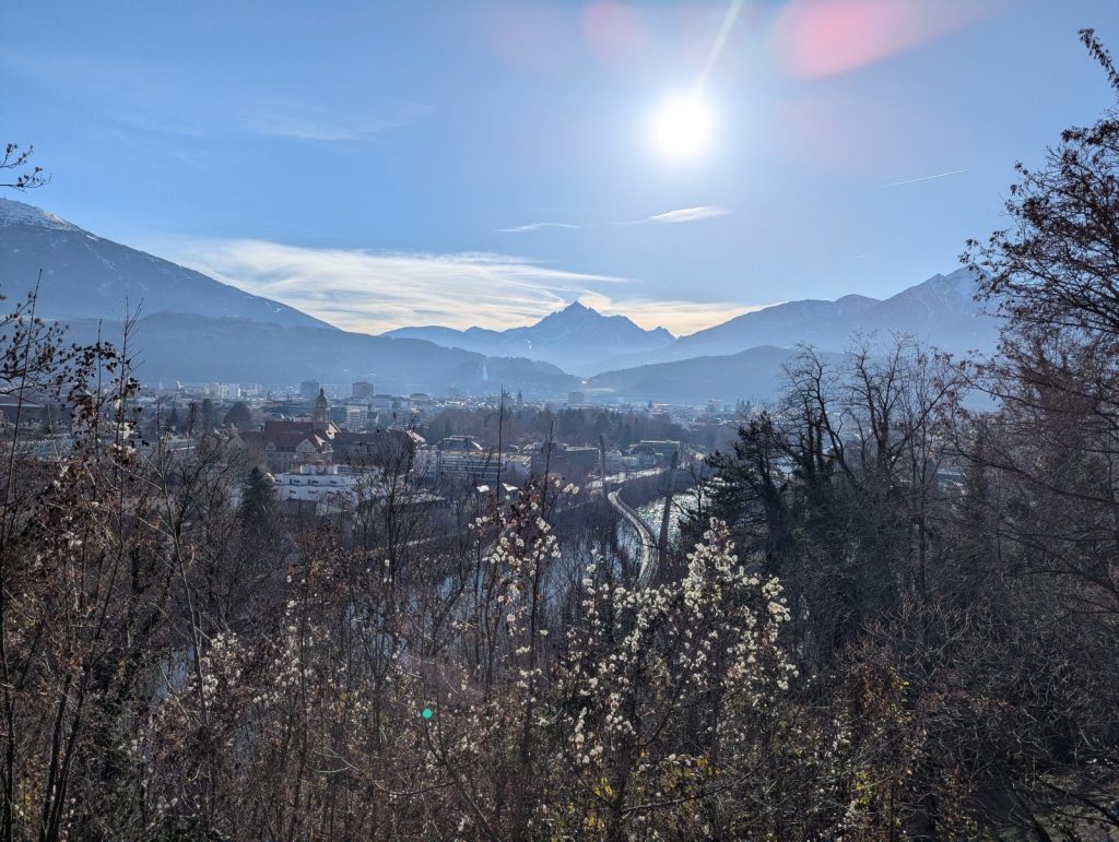 Sunlit panoramic view over Innsbruck and the Inn Valley from a hillside near the Alpenzoo, with layered Alpine peaks in the distance under a clear blue sky.