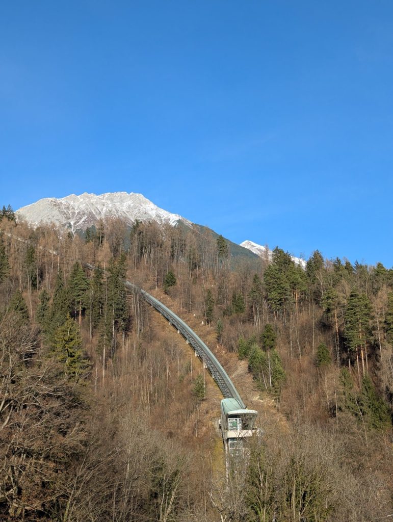Cable car line climbing a forested hillside in Innsbruck, with a snow-dusted mountain peak and clear blue sky in the background.