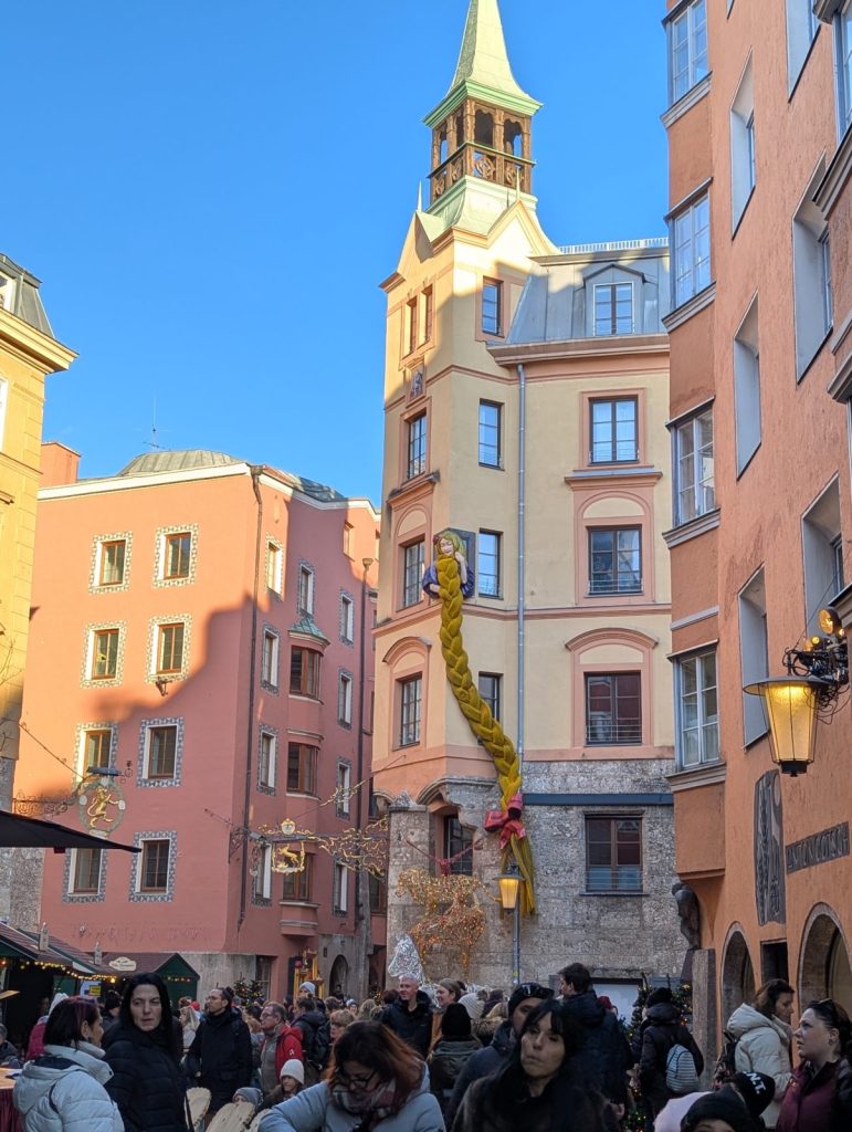 Crowded pedestrian street in Innsbruck Old Town lined with pastel buildings, looking towards the Stadtturm (City Tower) with a green dome in the distance.