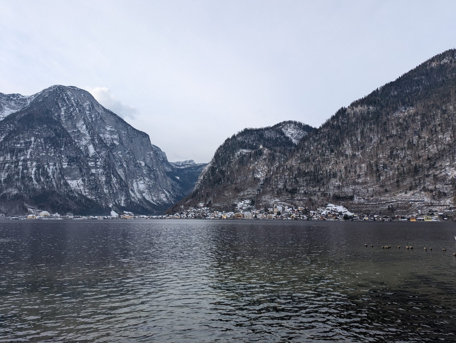 Winter view across Lake Hallstatt, with dark, rugged mountains rising behind the calm water under a pale, overcast sky.