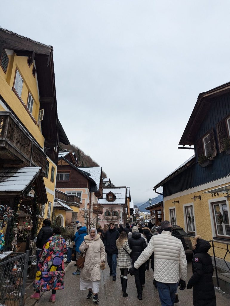 Busy narrow street in Hallstatt, with colourful historic houses, shop displays, and winter visitors walking uphill between the buildings.