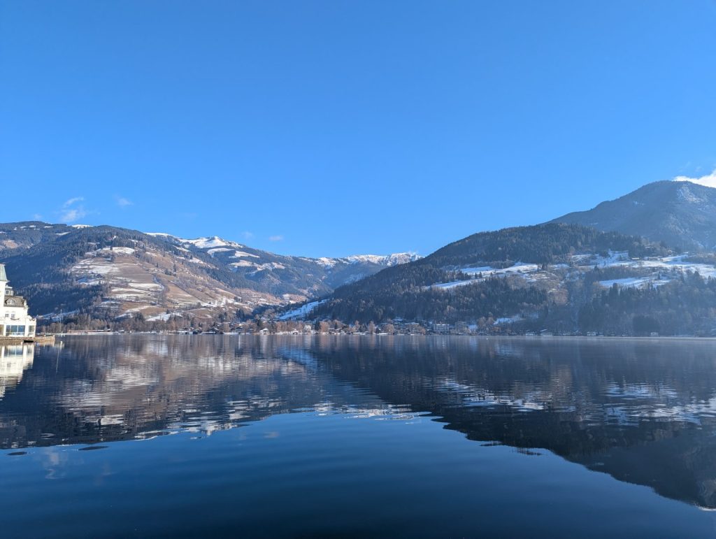 Panoramic winter view across Lake Zell in Zell am See, with glassy water reflecting snow-dusted mountains beneath a clear blue sky.