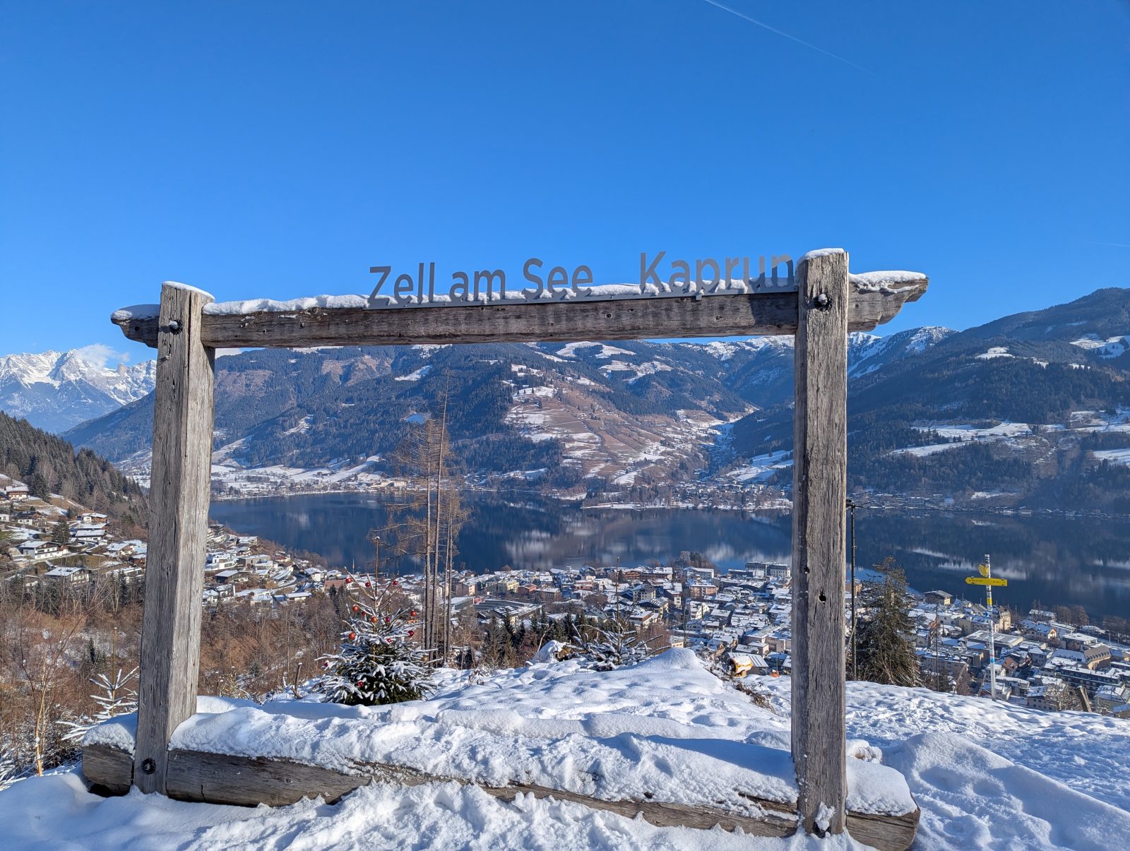 Wooden photo frame viewpoint sign above Zell am See, with snowy ground and panoramic winter views of the lake and surrounding mountains under a clear blue sky.