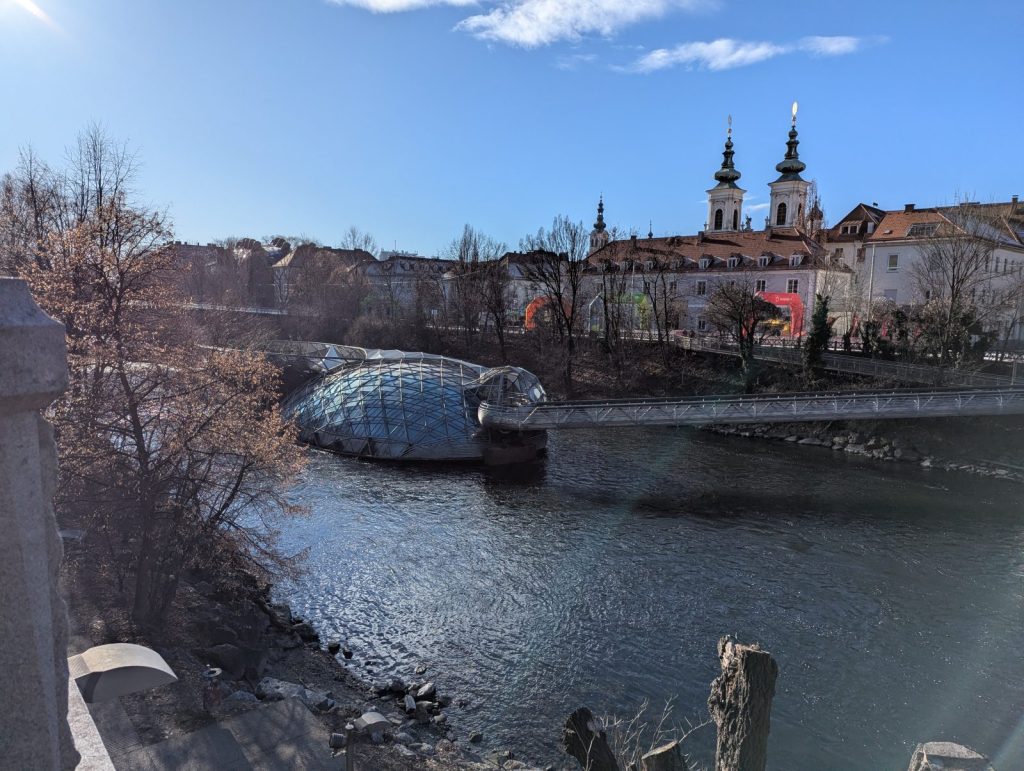View over the Mur River in Graz, with the Murinsel floating platform in the water and church towers rising in the background, framed by bare trees and historic buildings under a pale blue sky.