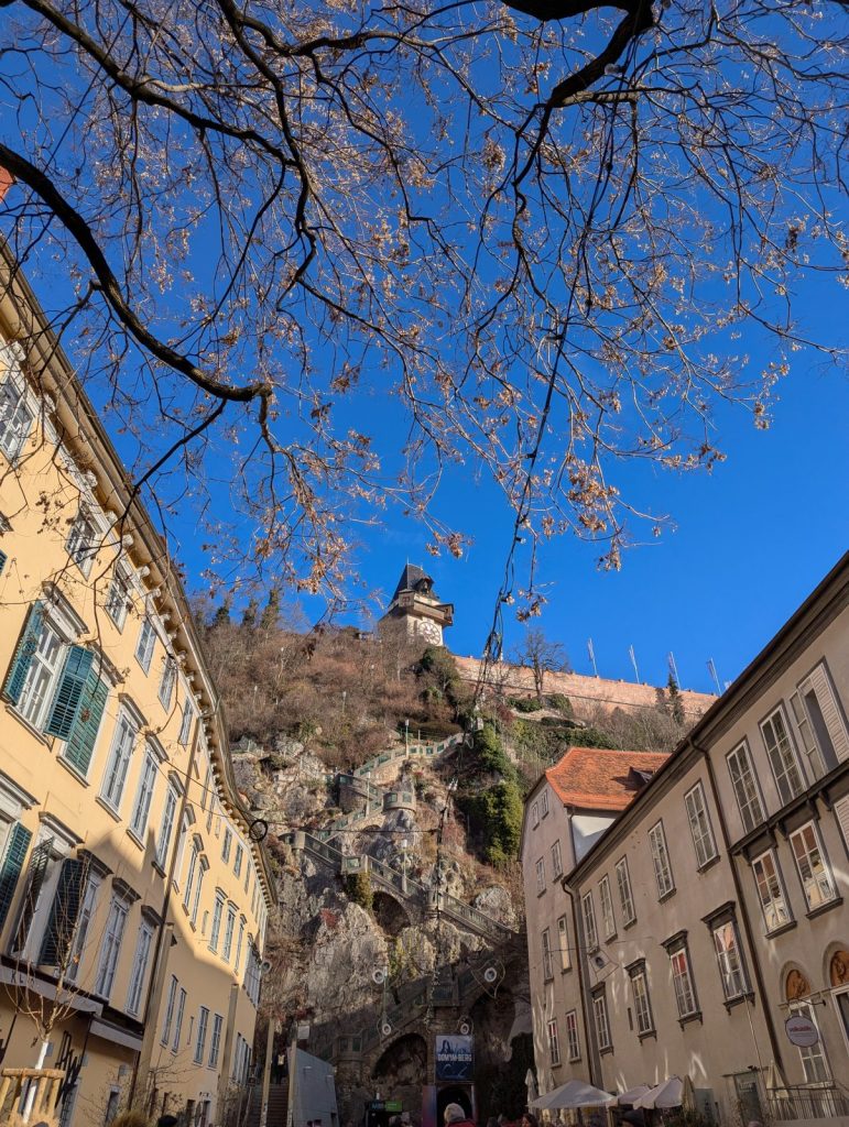 View up a narrow street in Graz toward the Schlossberg, with the Uhrturm perched on the rocky hilltop, framed by historic buildings, bare tree branches, and a clear blue sky.