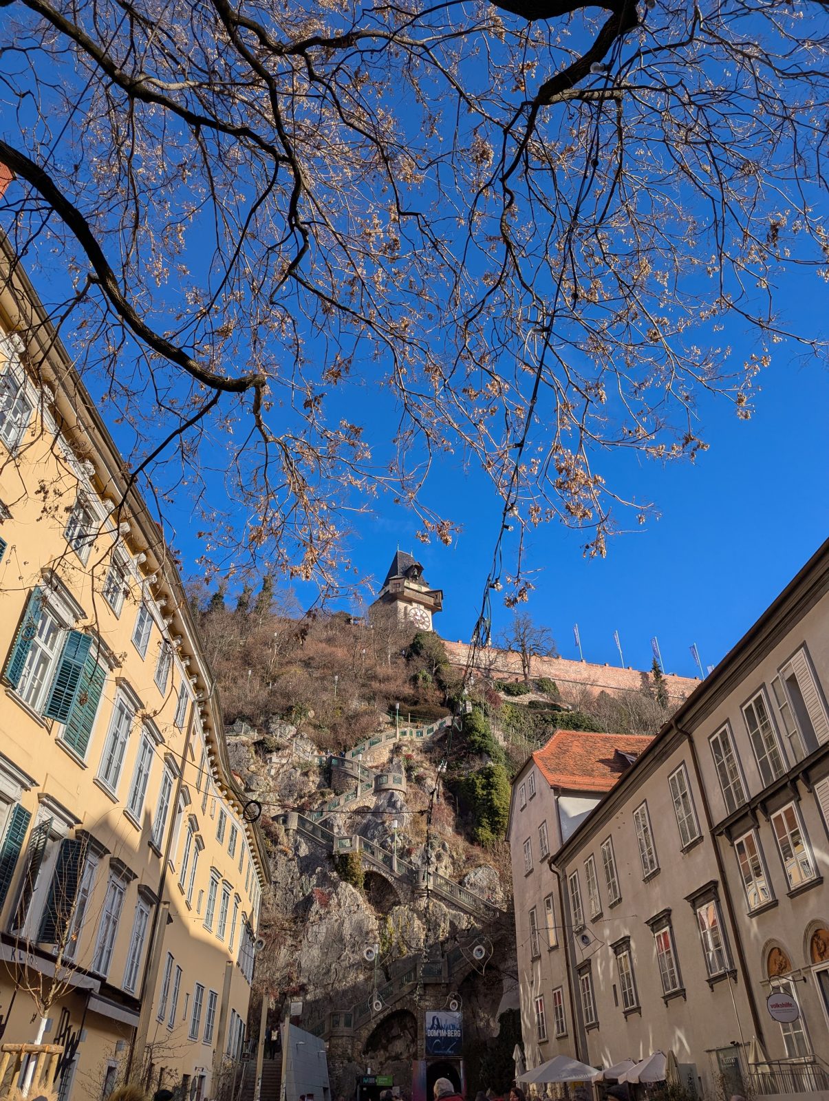 View up a narrow street in Graz toward the Schlossberg, with the Uhrturm perched on the rocky hilltop, framed by historic buildings, bare tree branches, and a clear blue sky.