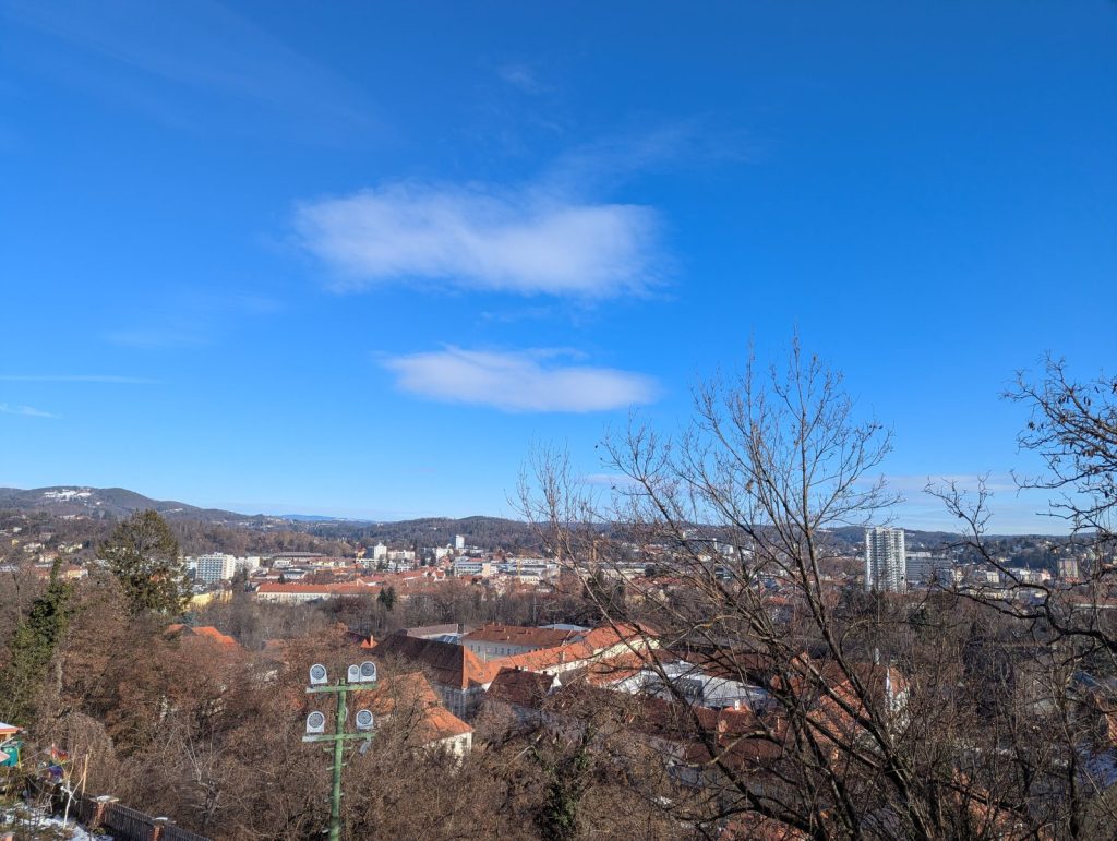 Panoramic view over Graz with red-tiled rooftops, church towers, and distant hills, seen from an elevated viewpoint with bare tree branches in the foreground under a bright blue sky.