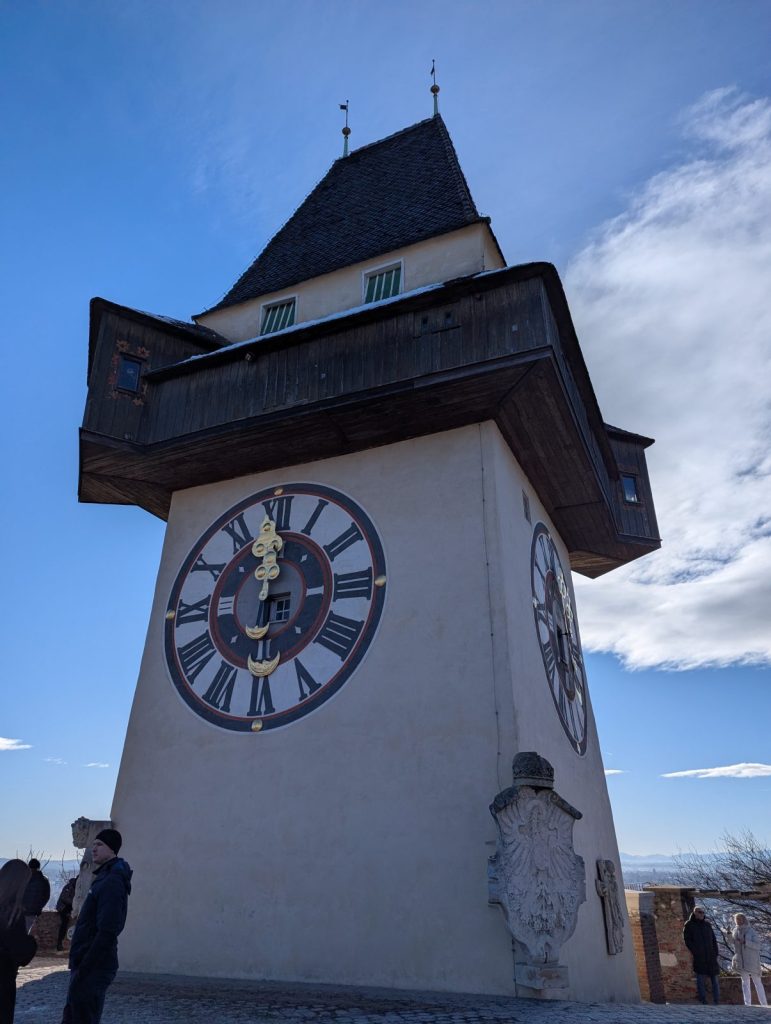 Graz Clock Tower (Uhrturm) viewed from below, showing the large clock face with Roman numerals on a white tower and the dark wooden roof against a partly cloudy blue sky.