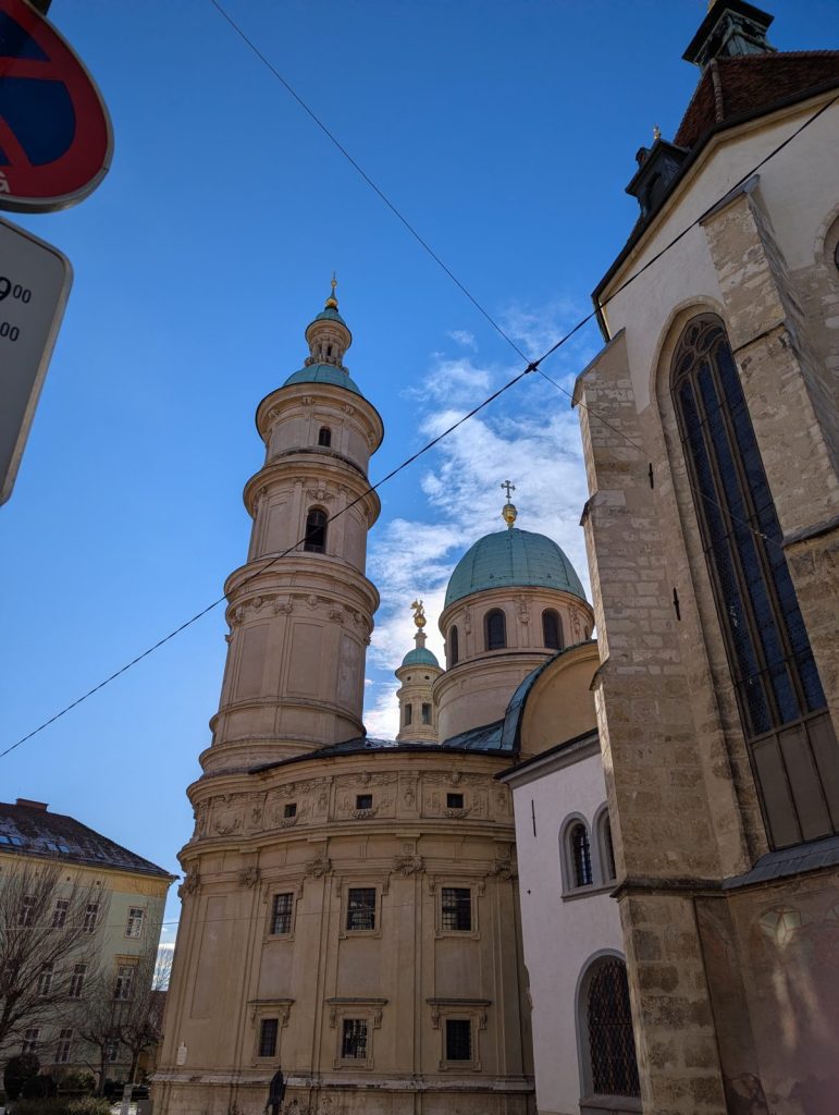 View looking up at Graz Cathedral and the Mausoleum of Emperor Ferdinand II, showing the tall stone tower, green domes, and church walls framed by narrow streets under a clear blue sky.